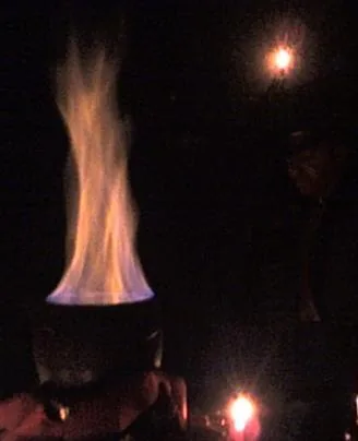 Chama elevando-se de um caldeirão num ambiente escuro iluminado por velas